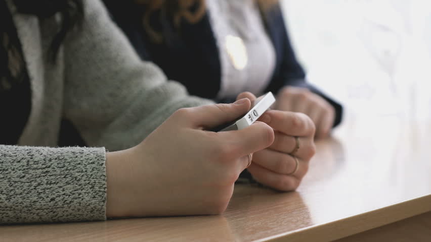 Schoolgirls dressed in cardigans sit at the school table at the school. One girl holds a white smart phone her hands. Close-up