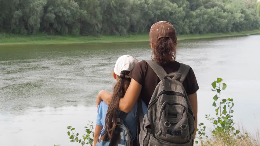 Mom and child are looking at the current river. A woman with a little girl embraced standing on the river bank.