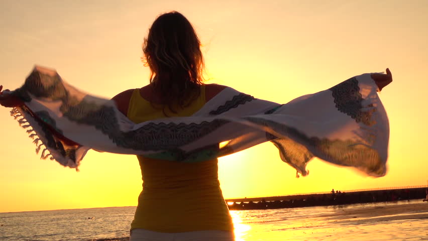 CLOSE UP, SLOW MOTION, SILHOUETTE: Young woman standing on beach looking at rippling ocean at sunset. Golden light sun setting behind the horizon. Girl on seashore holding scarf waving in summer wind