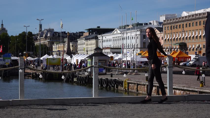 Young woman in black come and stay at Helsinki port embankment, market square on background. Tourist lady admire beautiful city at warm sunny autumn day. Many tents and stalls at weekend bazaar