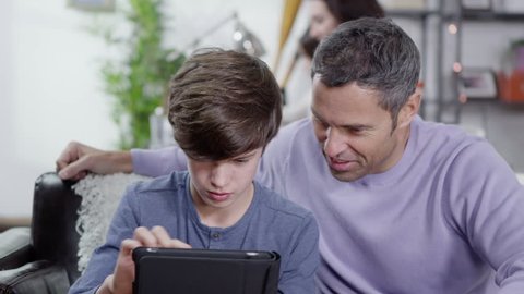 A father and son are browsing the internet on a digital tablet whilst mother and daughter can be seen in the background Stockvideo
