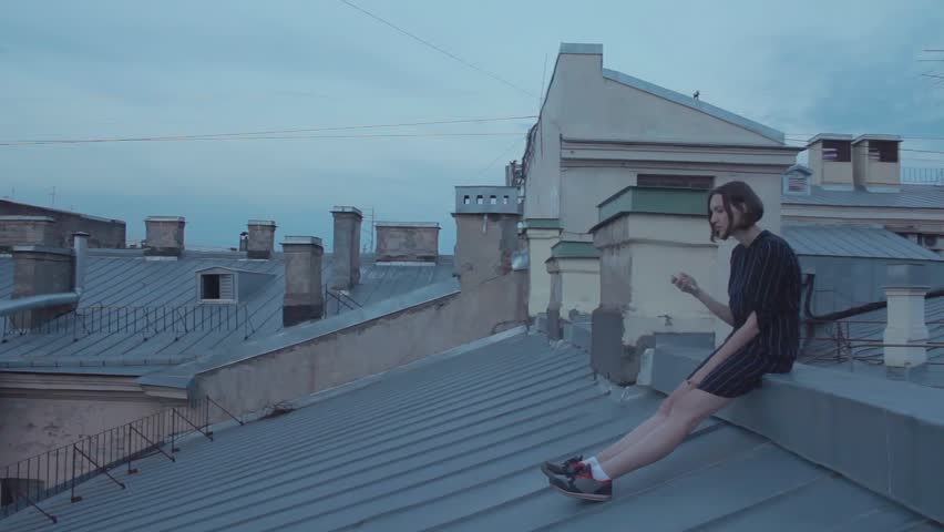 Young girl using the smartphone sitting on the roof. Girl with short hair, wearing a dark blue dress with stripes is on the roof building in old centre of city
