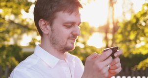 Portrait of Young Man Using Smart Phone in a Sunny Park. 4K SLOW MOTION, DCi. Handsome man, using cellphone outdoors, Lens Flare. Technology in everyday life.