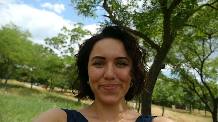 A cheerful young brunette girl smiles happily and spins left and right in a picturesque park with a lot of trees and a nice lawn on a sunny day in summer in slow motion