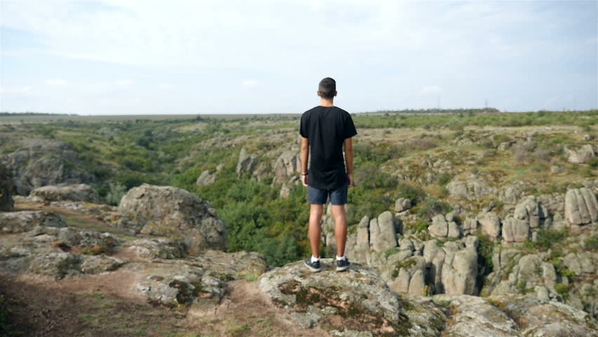 A striking view of a young man, dressed in black shirts and a t-shirt, while standing on a top of a cliff and raising his hands near a deep abyss on a picturesque hill in summer in slow motion.