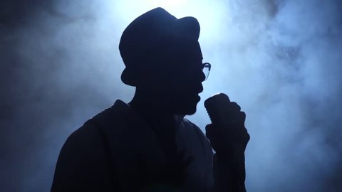 African American man comes out of the smoke to the microphone singing in a dark recording studio. Black background. Silhouette. Slow motion. Close up