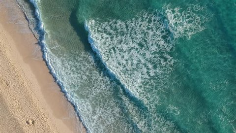 Aerial drone view of a beach near Corralejo on the island of Fuerteventura, Spain. Rolling waves gently breaking onto the sand. Sand texture visible on ocean floor.