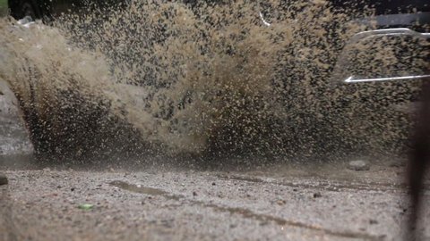 SLOW MOTION CLOSE UP: Detail of a SUV car tyre driving over deep puddle and splashing water across the dirt road. Black SUV car tires driving on wet dirt road after rain and over muddy puddle.