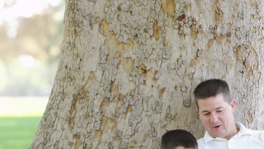 Father and son sit under a tree, playing with a digital tablet.