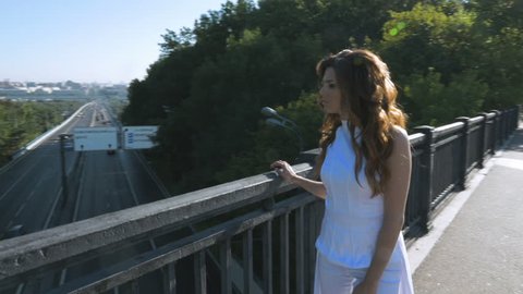 A young woman with long hair walks along the pedestrian bridge. She looks at the cars that are driving downstairs.