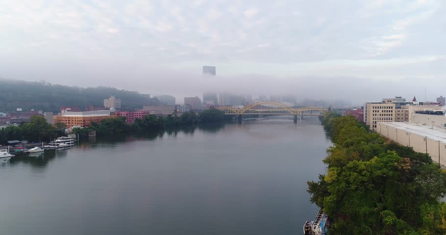 An early morning foggy aerial establishing shot of the Pittsburgh skyline with the Steel Building poking up above the fog. The Allegheny River is in the foreground.  	