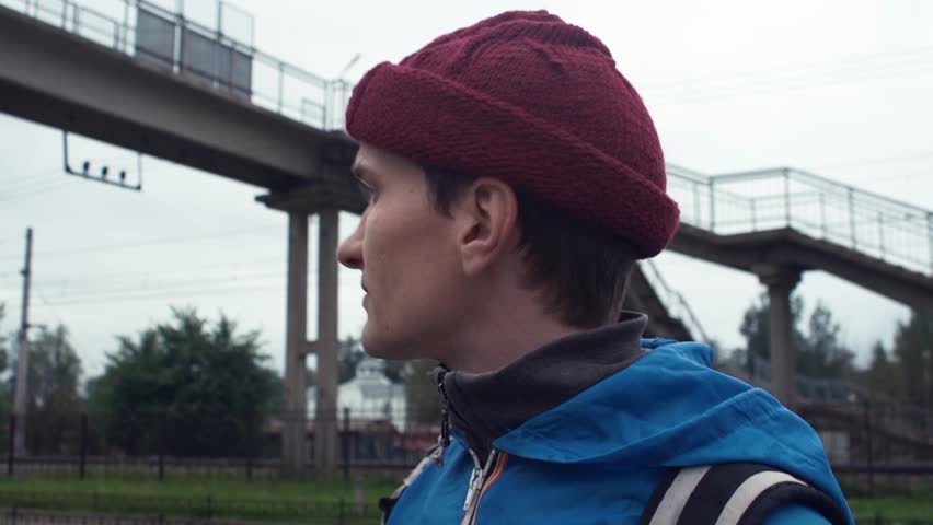 Young hiker dressed in blue jacket and knitted hat with backpack standing on pedestrian bridge over railway looking far away. Freedom travel adventure theme | Shutterstock HD Video #30812632