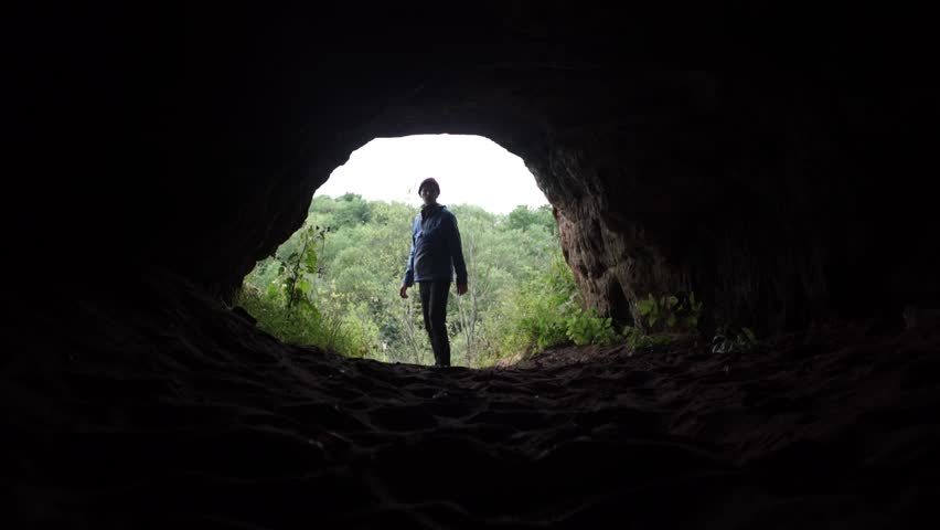Young explorer enters to dark cave chamber under low stone vault and walk into darkness. Traveler inside the cave. A look from the cave. Deep forest at background. Royalty-Free Stock Footage #30812815