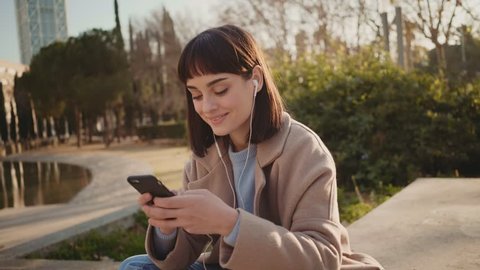 Young attractive cute millennial hipster woman chats and flirts on messenger application on smartphone while sitting outside in park in winter,concept social media, sexting and communication