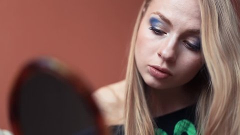 Attractive blonde caucasian woman doing her make up puts on blue eye shadows looking into small handheld mirror