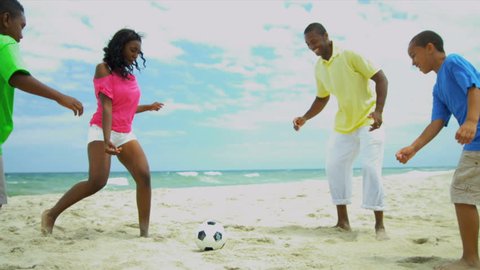 African American children enjoying summer kicking football with father on beach