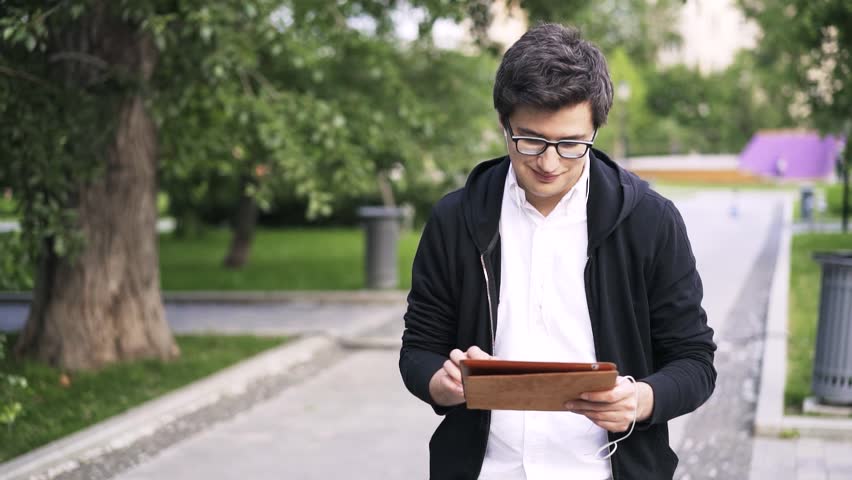 Young man wearing glasses and a black hoodie is listening to the music, using his tablet and walking in a park. Handheld slow motion medium shot Royalty-Free Stock Footage #30830314