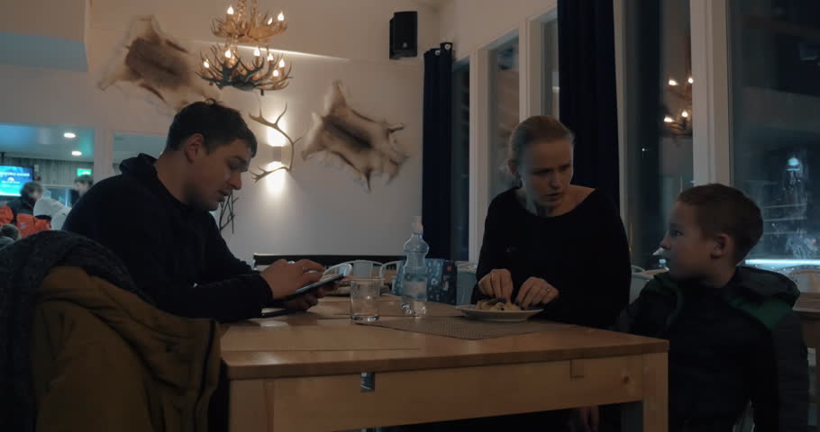Family having meal in Finnish cafe in winter evening. Mother and child eating while father taking a mobile photo of a kid. Interior with fur and antlers