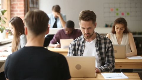 Young businessman sitting at desk using laptop in coworking space with multi-ethnic people, startupper developer working distantly in shared office with diverse coworkers, looking at camera smiling