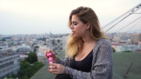 Beautiful sad young blonde sitting on the roof high above the metropolis and blowing soap bubbles