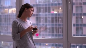 Young woman standing by window drinks coffee HD slow-motion video. Girl looking at morning or evening city landscape at home flat apartment