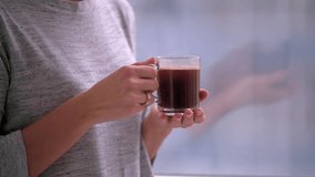 Young woman drinks coffee or tea by window HD slow-motion video. Close-up of girl hand holds cap. Morning or evening at home flat apartment
