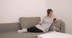 Woman on home sofa using smartphone and drinks water 4k video. Girl online in mobile phone at modern flat apartment 