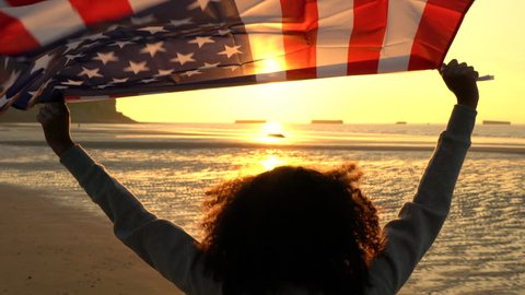 4K video clip of mixed race African American girl teenager female young woman holding an American US Stars and Stripes flag on a beach at sunset or sunrise Stockvideó