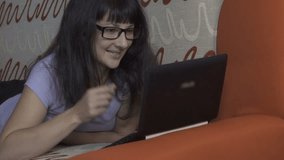 Girl student studying on laptop. Beautiful young woman with glasses video chatting on her laptop computer while lies on the couch in the living room. The girl sends an air kiss.