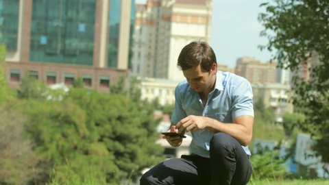Young man sitting on grass with smartphone in the park with green tress and business buildings background