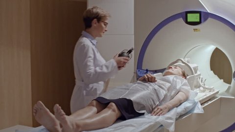 PAN of female medical technician in glasses putting headphones on young woman lying on patient table of MRI machine, then performing scan