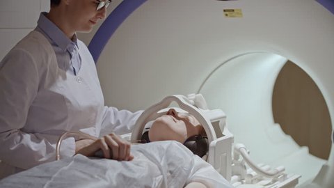 Tracking of cheerful female radiologist putting headphones on woman in disposable gown lying on patient table, then moving her into MRI machine for scan