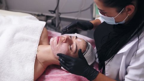 Young woman with blemish skin lying on the couch while professional cosmetologist is applying face cream and making face massage in spa salon