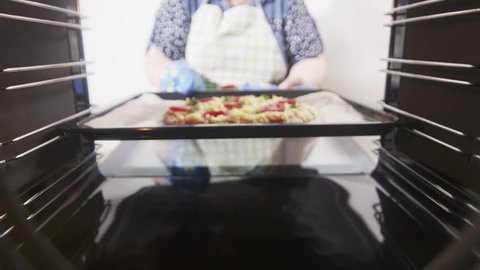 View from inside of the oven. Cooking italian pizza in electric convection oven. Woman placing pizza on a baking sheet inside hot cooker. Frozen pizza topped with salami tomatoes mozzarella cheese.