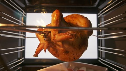 Cooking roasted whole chicken on the rotisserie spit in hot convection oven. Browned chicken rotates during broiling under a grill element. Meat juices dripping onto a baking tray. Inside view.