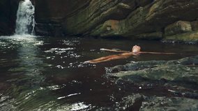 Woman floating and relaxing inside river by a waterfall