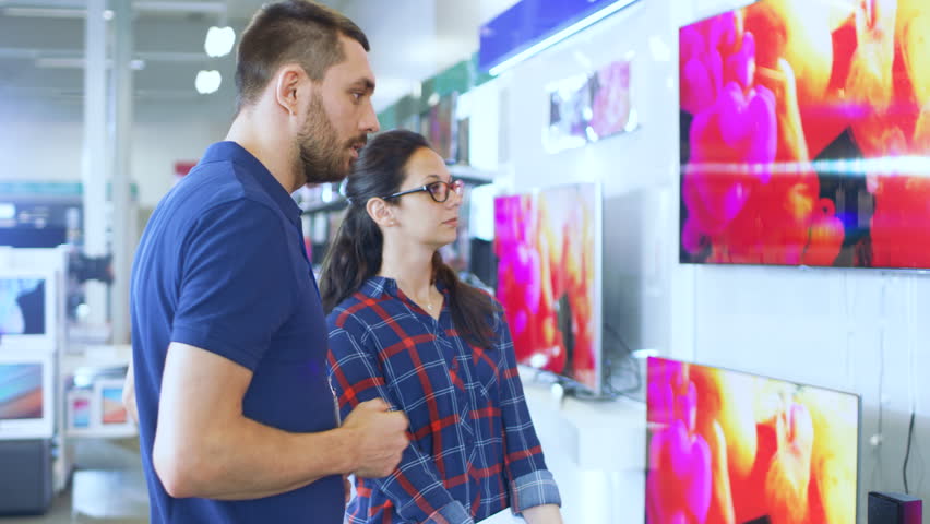 In the Electronics Store Professional Consultant Provides Expert Advice to a Young Woman Who Looks for a  New 4K UHD TV to Buy. In This Modern Store all the Latest TV Models Are Hanging on the Wall. Royalty-Free Stock Footage #30883903