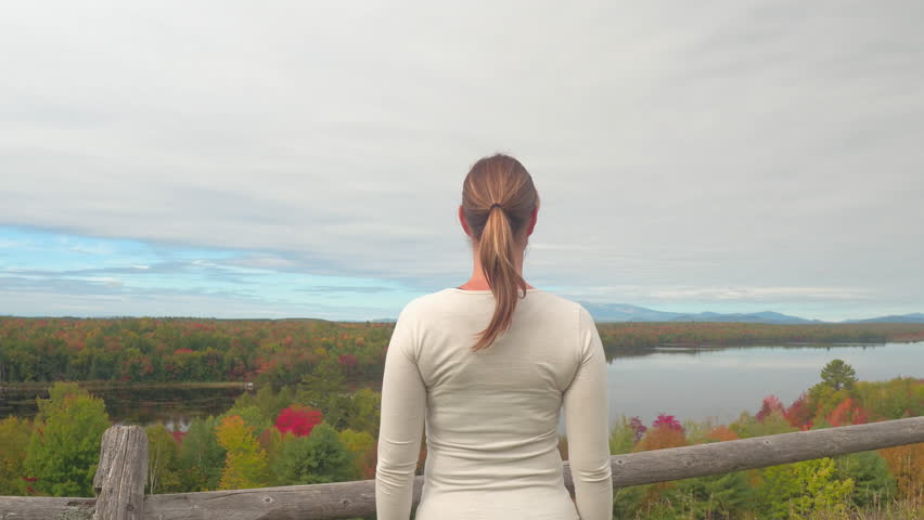 Young brunette woman outstretching arms proudly overlooking stunning autumn forest reflecting in glassy lake. Cheerful girl raising arms in success on picturesque viewpoint of colorful autumn nature