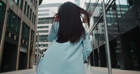 epic Steadicam shot of a young beautiful Asian girl with smartphone going to work around office buildings, back view, flowing hair