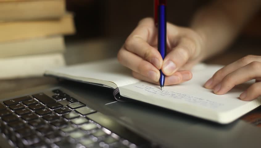 Close-up of a young woman writes a pen in a notebook (HD), laptop and books in