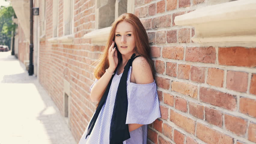 Happy, beautiful woman with red hair talking on cellphone by the wall in the city
