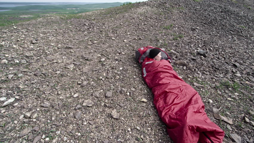 High angle of Asian woman napping in sleeping bag during solo hiking trip in mountains