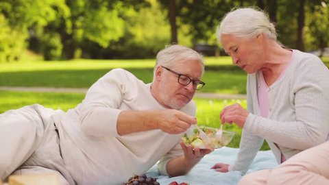 old age, leisure and food concept - happy senior couple eating salad at picnic in summer park