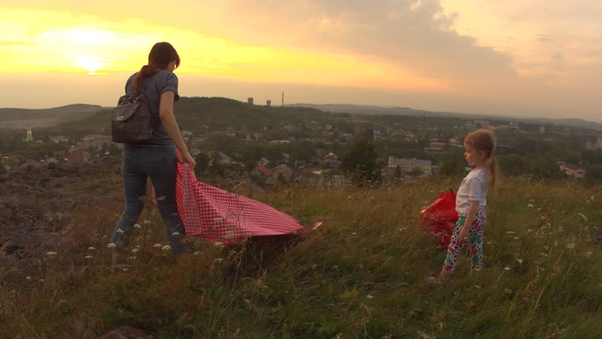 A young woman setting up red checkered blanket for picnic, little girl helps her. Young family holds a weekend outdoors together on a hill in the background of the urban landscape.