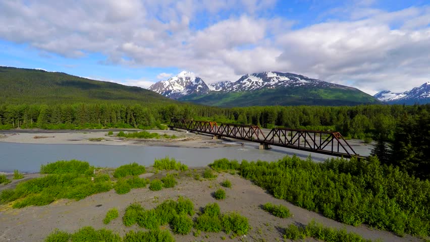 Alaska 4k Drone footage tracking over old iron railroad bridge with glacier mountain in background then going over bridge and down a river bed