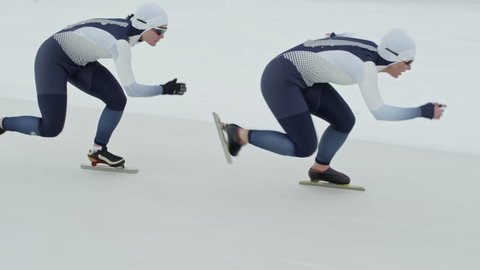 Tracking with slow motion of professional female speed skaters wearing spandex full-body covering suits sprinting along track in ice rink