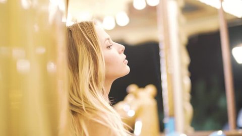 attractive blonde girl looks at the camera, smiling, fooling around. With carousel on background. Slow motion
