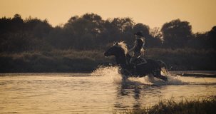 Slow motion 240fps shot.Woman wearing cowboy outfit riding horse on the background of beautiful sunset and water. Girl model on horseback on river water at sunset, backlit with sunshine.
