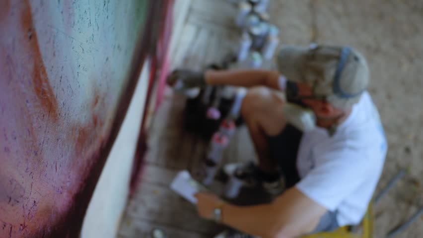 Graffiti artist in protective mask and gloves painting with red aerosol spray on the street wall. Slow motion.