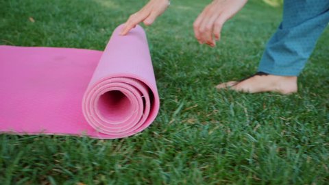 Young woman doing yoga exercise - opening her pink yoga mat on green grass at the park. Slow motion.